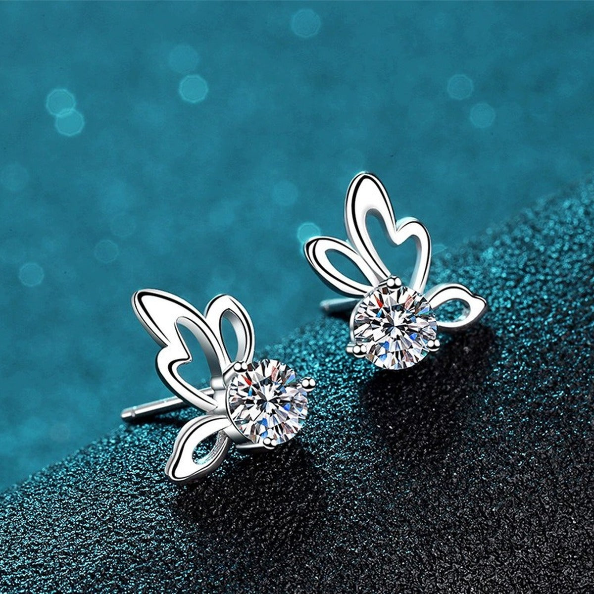 Butterfly Moissanite Stud Earrings in Rhodium-Plated S925 Sterling Silver on blue-green fabric showing angled setting view
