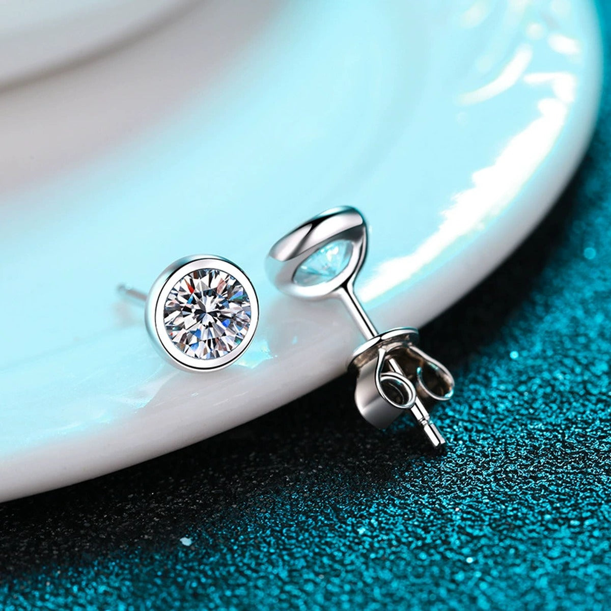 Bezel Round Moissanite Stud Earrings in Rhodium-Plated S925 Sterling Silver on blue-green fabric showing angled setting view