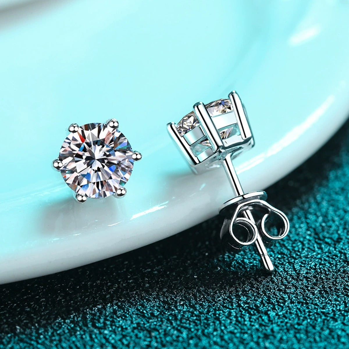 Classic Round Moissanite Stud Earrings in Rhodium-Plated S925 Sterling Silver on blue-green fabric showing angled setting view