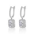 A pair of silver-toned drop earrings featuring a rectangular halo setting with a large clear emerald-cut center stone and smaller surrounding stones, suspended from huggie hoops with a single row of small stones, on a white background with a soft reflection below.