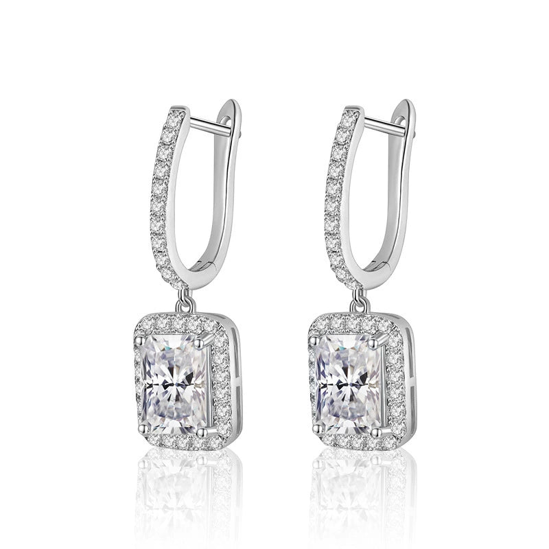 A pair of silver-toned drop earrings featuring a rectangular halo setting with a large clear emerald-cut center stone and smaller surrounding stones, suspended from huggie hoops with a single row of small stones, on a white background with a soft reflection below.