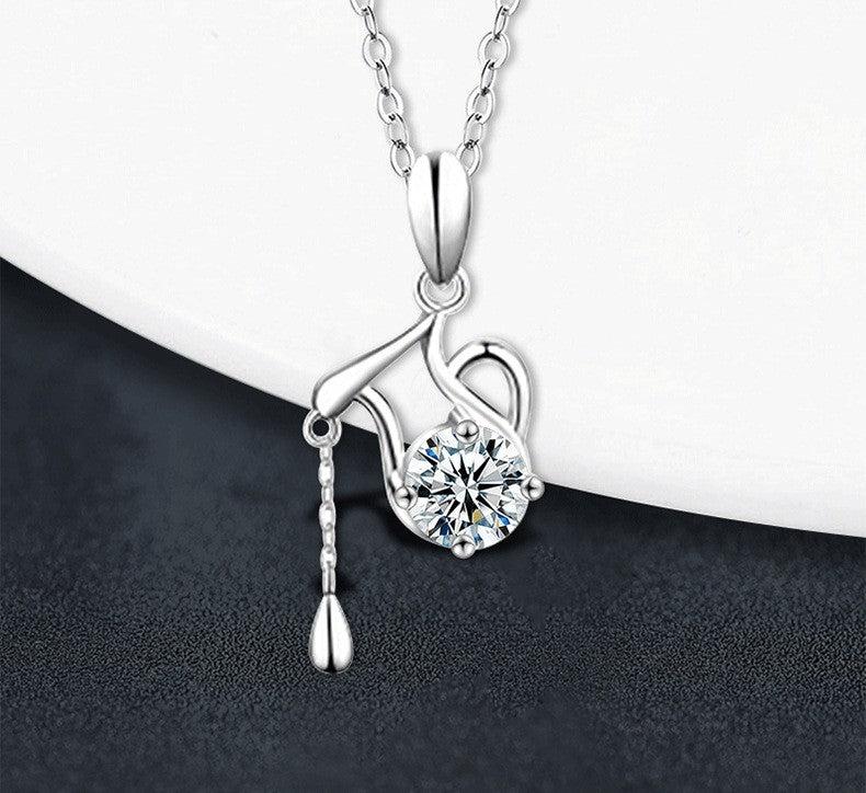 A silver pendant necklace featuring a teapot design with a round clear gemstone and a dangling teardrop charm, displayed on a white curved surface against a dark textured background.