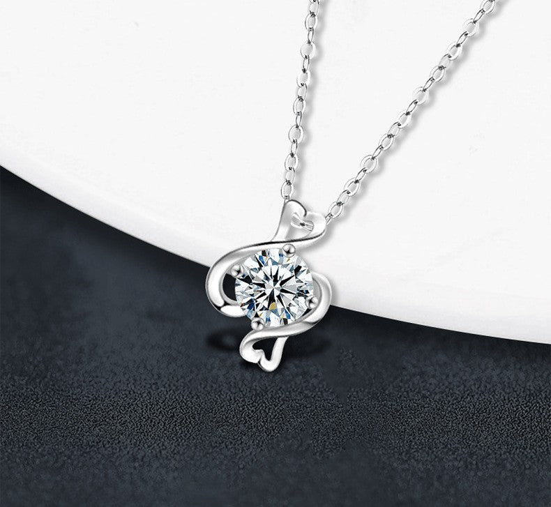A silver necklace with a round clear gemstone pendant, featuring a swirling openwork silver design and a small heart-shaped accent, displayed on a white curved surface against a dark textured background.
