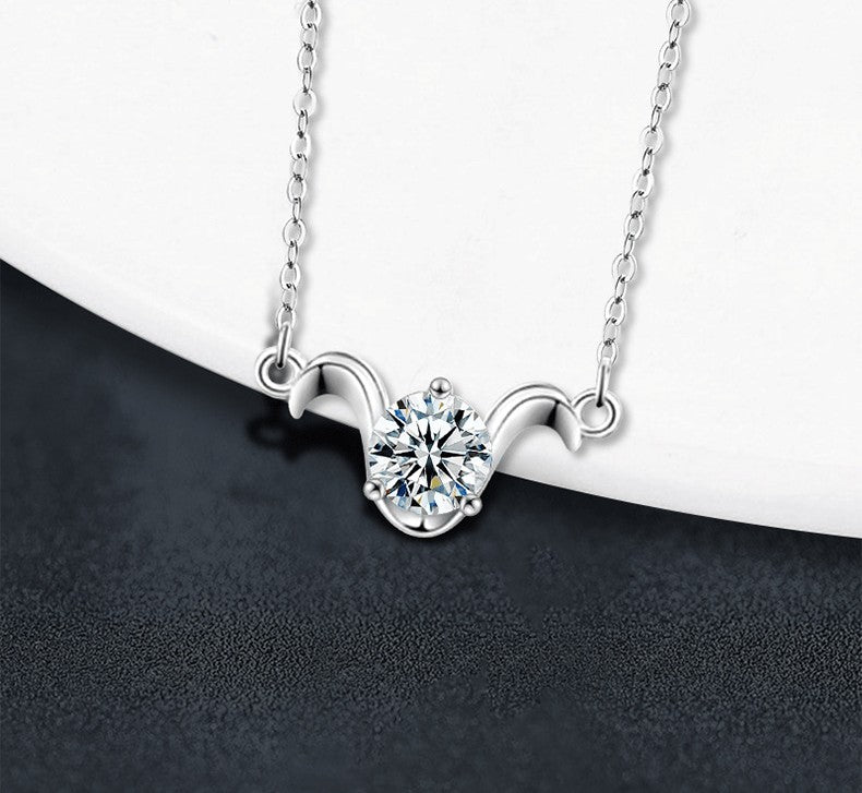 A silver-toned necklace with a delicate chain and a curved pendant setting holding a clear round gemstone, displayed on a white curved surface against a dark textured background.