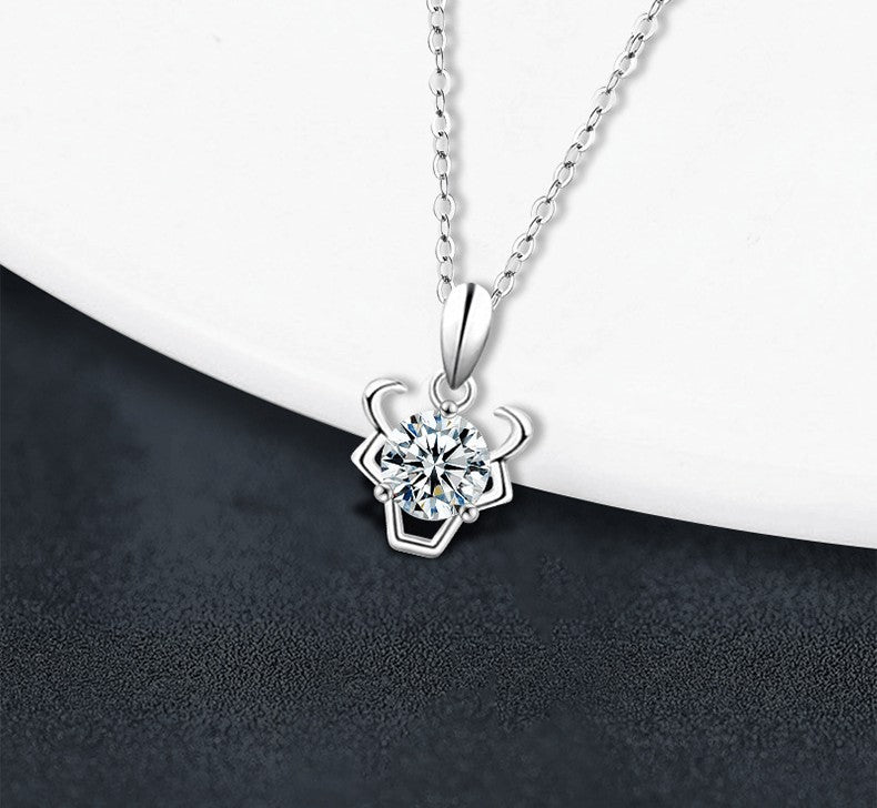 A silver-tone necklace with a flower-shaped pendant featuring a clear round gemstone, displayed on a white curved surface against a dark textured background.