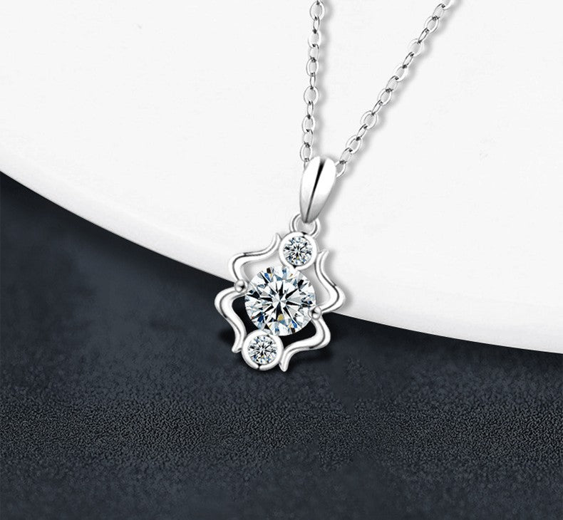 A silver pendant necklace with a floral design featuring a central round clear gemstone and three smaller accent stones, displayed on a white curved surface against a dark textured background.